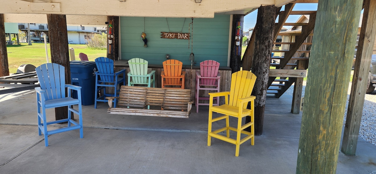 A covered outdoor area features a mix of colorful adirondack chairs in shades of blue, orange, pink, and yellow. A wooden swing is positioned among the seating, providing a relaxed space. The setting is complemented by sturdy wooden support beams and a glimpse of the grassy exterior.
