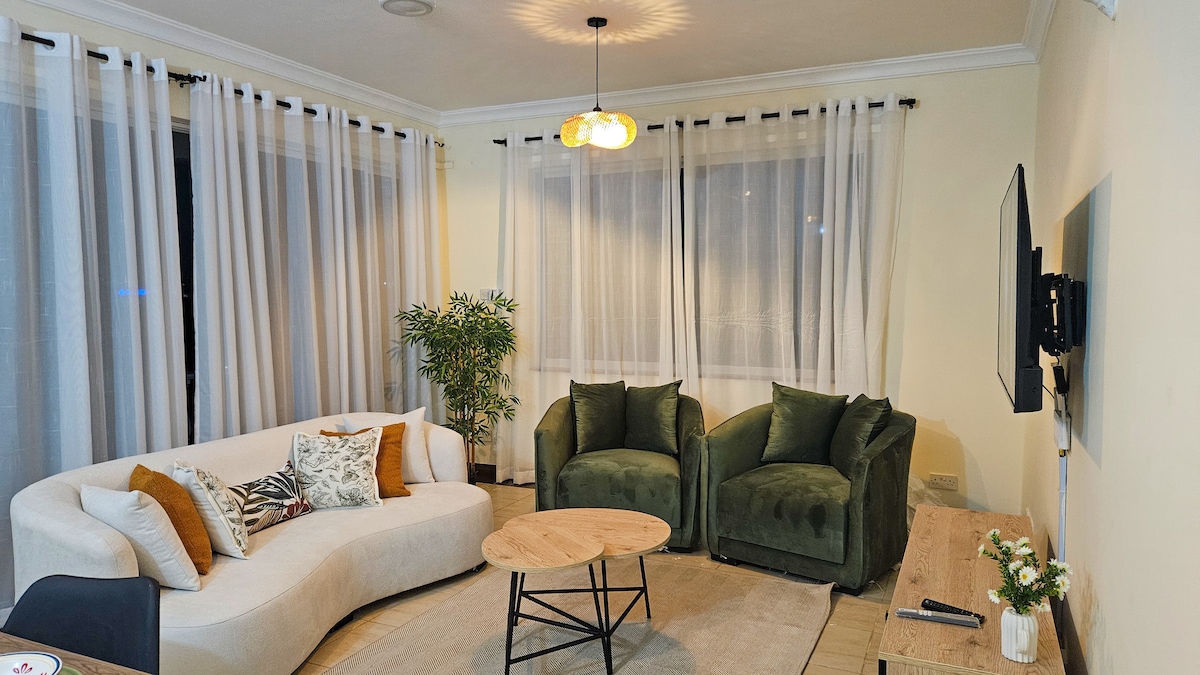 The living room features a large curved sectional sofa adorned with decorative cushions. Two green armchairs complement a round wooden coffee table at the center. Soft drapes frame the windows, allowing natural light to fill the space, enhancing the inviting atmosphere.