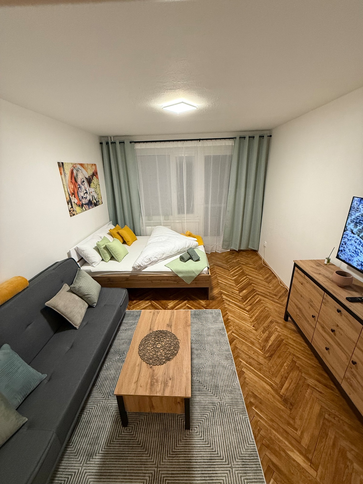 A bright living space features a spacious bed with light bedding, complemented by colorful accent pillows. A gray sofa provides additional seating, while a wooden coffee table sits atop a patterned rug. Natural light filters through sheer curtains, enhancing the cozy atmosphere.