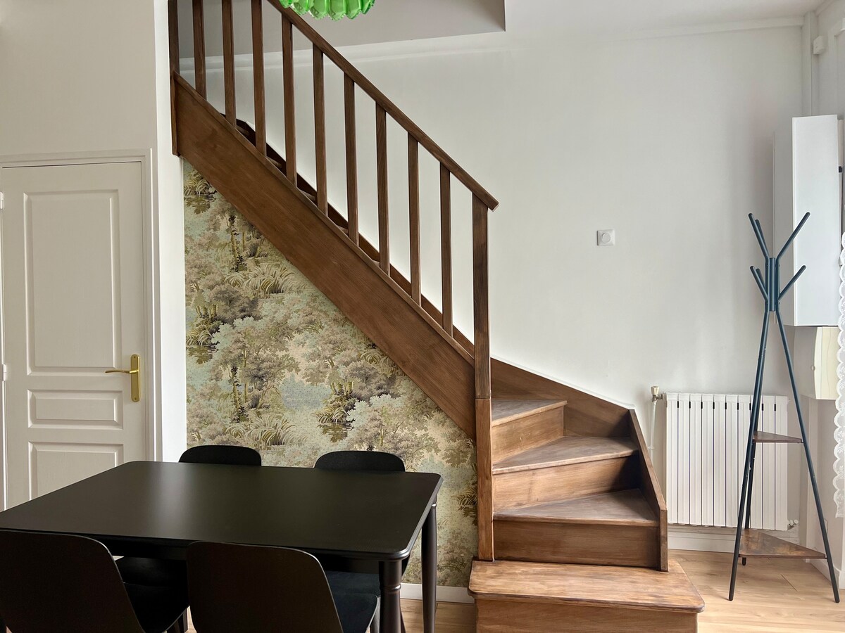 A wooden staircase with a simple railing leads to the upper level. A floral-patterned wallpaper adds a touch of design to the wall. Below, a black dining table with four chairs is positioned, providing a functional space for meals.