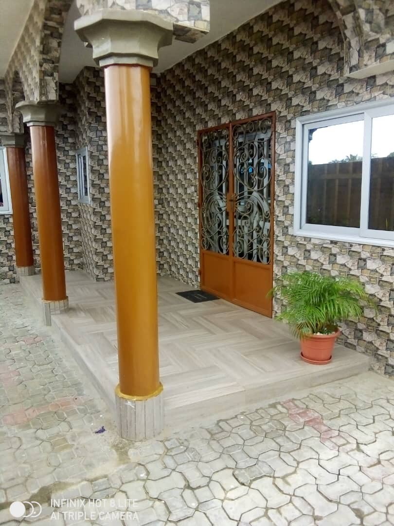 An exterior view of the entrance features a decorative glass door framed by two tall pillars. The entrance is set on a textured tiled surface with a potted plant placed near the door. A patterned stone wall adds to the visual interest.
