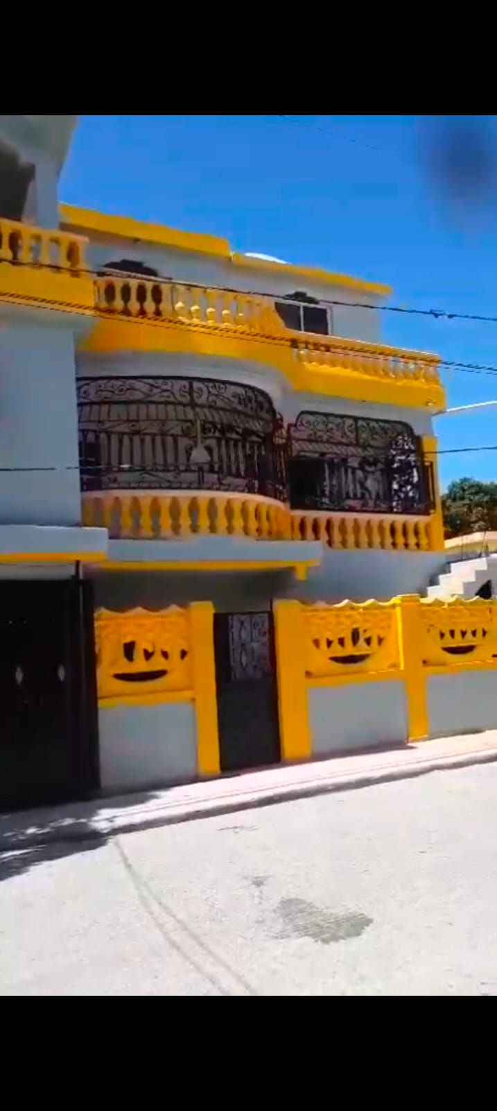 The vibrant exterior of a two-story building is presented, featuring bold yellow accents and decorative black wrought iron railings. A unique façade showcases intricate artwork, while a staircase leads to the upper level. A cheerful, sunny street view enhances the lively character of the home.