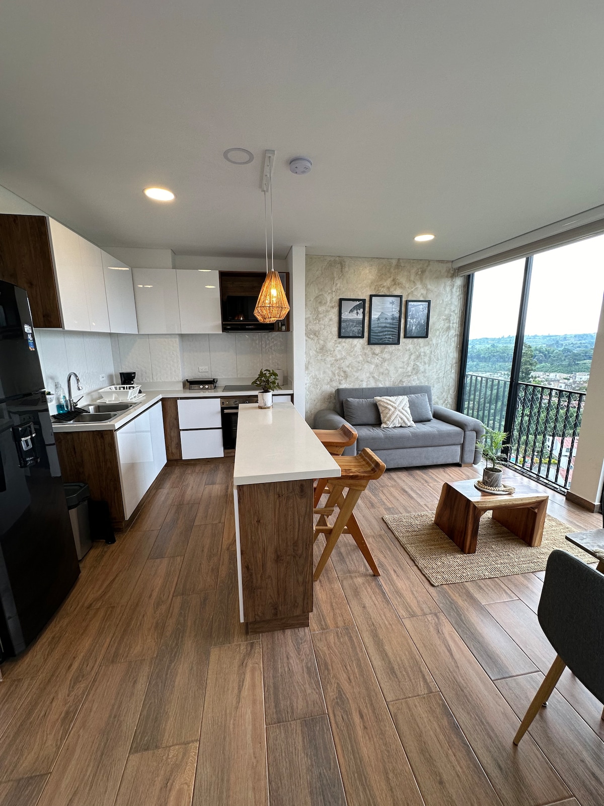 A modern kitchen is combined with a cozy living area. Neutral-toned cabinets and a dark refrigerator complement the light countertops. A small dining table is positioned near the window. Natural light floods in, showcasing the inviting seating area and scenic views of the surrounding landscape.