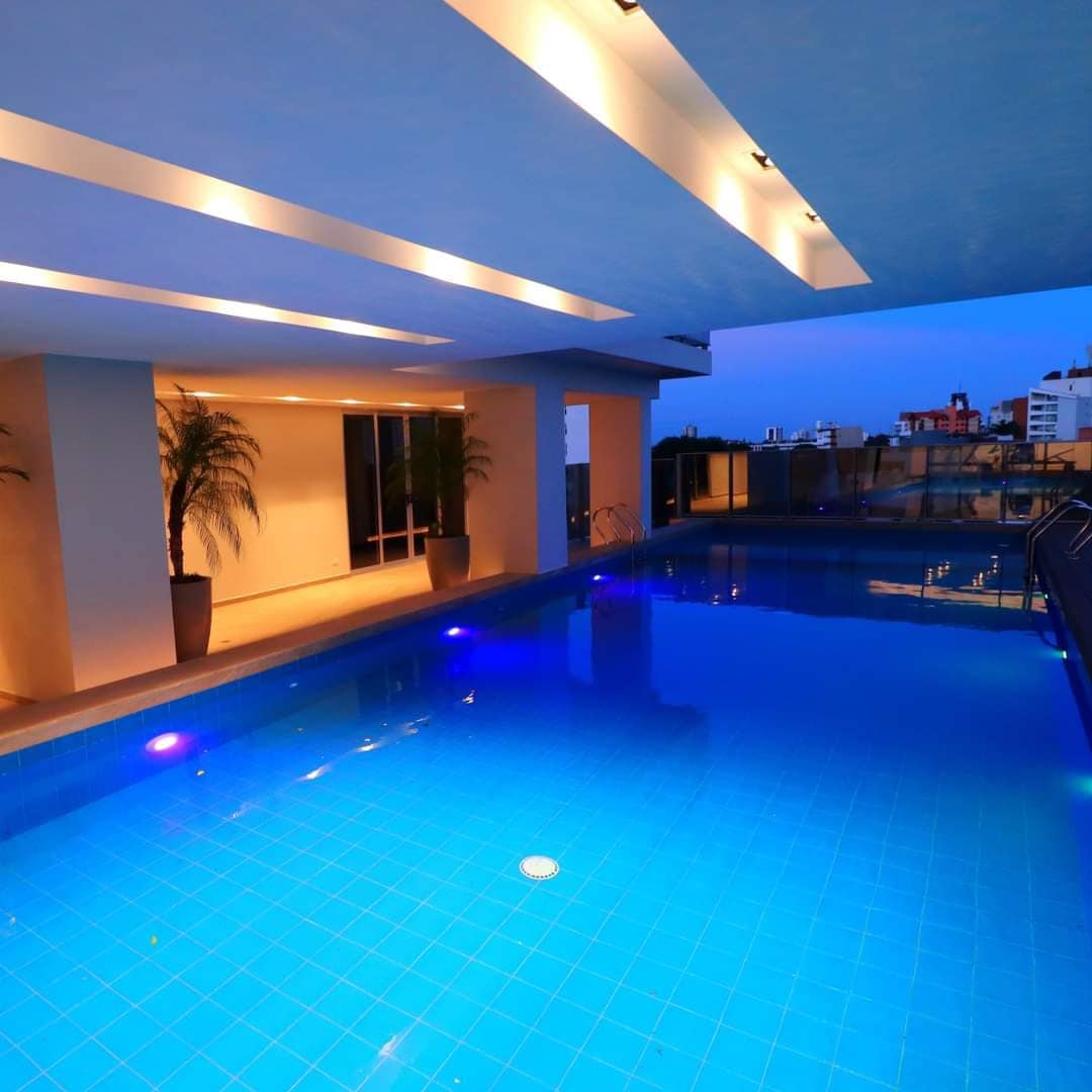 An inviting infinity pool is surrounded by modern architecture, with gentle lighting illuminating the water's vibrant blue hue. Tall indoor plants in decorative pots enhance the atmosphere, while views of the city skyline can be seen in the background as dusk settles in.
