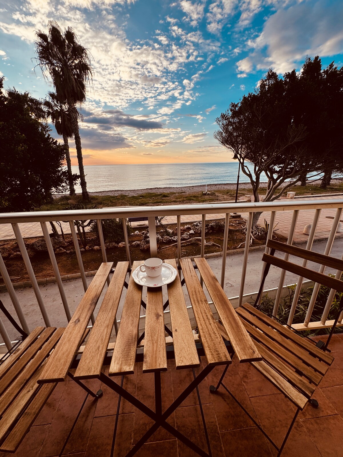 A wooden bistro table with two folding chairs is set on the balcony, facing the ocean. A cup sits atop the table, complemented by a calming view of the beach framed by palm trees. The colorful sky transitions from warm hues to blue above the horizon.