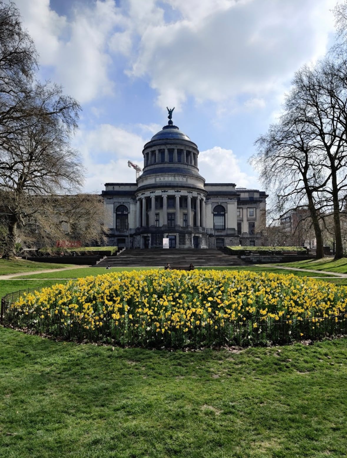A grand architectural building is set against a clear sky, featuring a prominent dome and adorned with a statue atop. Surrounding the structure, a vibrant display of yellow flowers blooms in a well-maintained garden, with green grass and tall trees framing the scene.