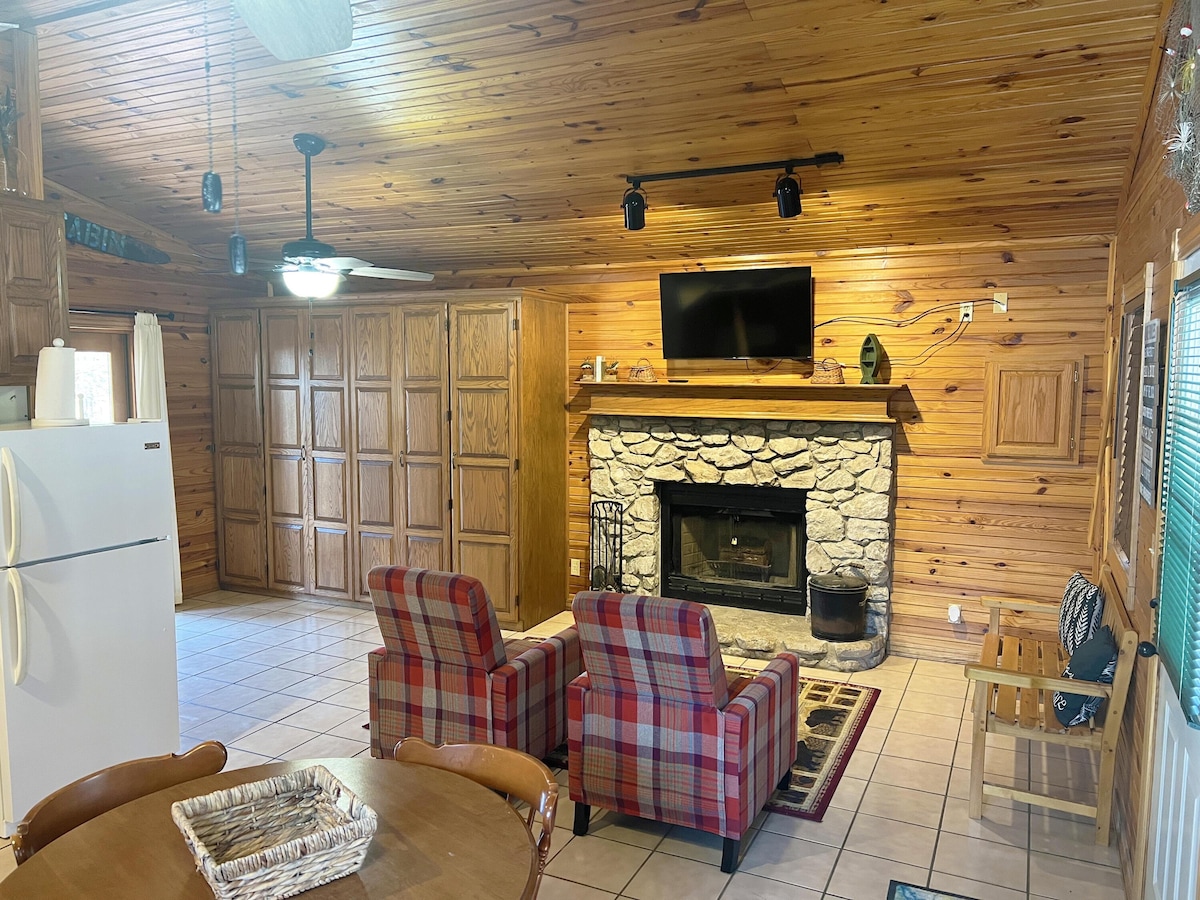 The cabin interior features wooden paneling and a stone fireplace as a focal point. Two plaid chairs are positioned in front of the fireplace, while a television hangs above. A kitchen area is visible, along with a round dining table, and ample storage cabinets line the walls.