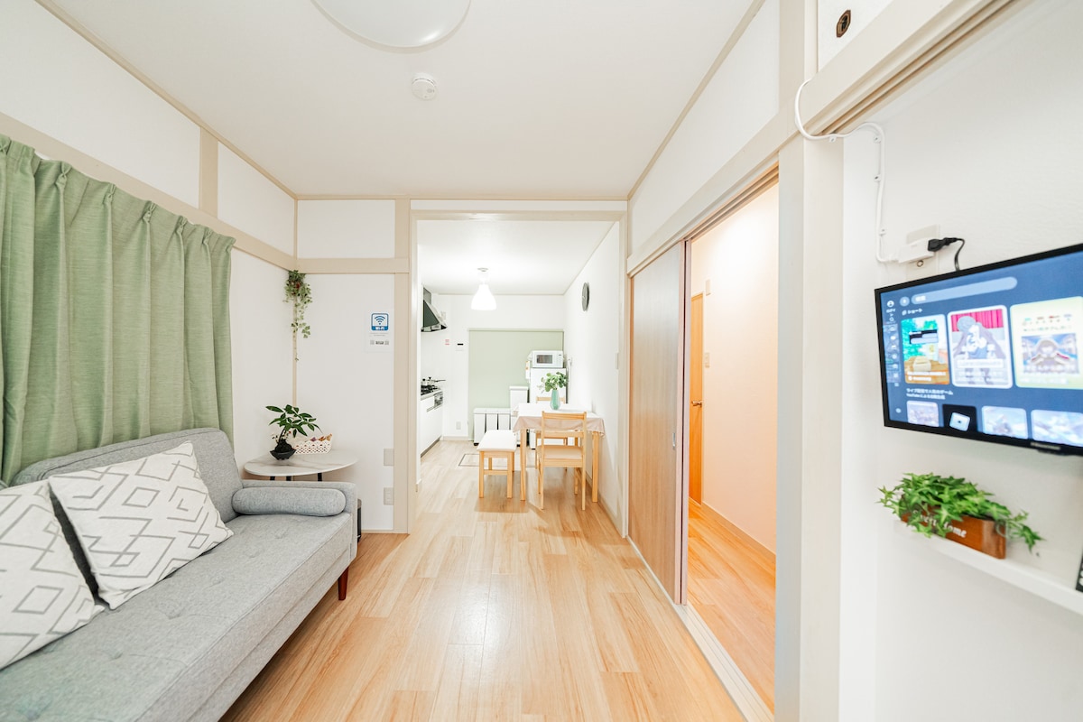 A welcoming living area features a light grey sofa with geometric patterned cushions. A small dining table with two chairs is positioned nearby, while a glimpse of the kitchen and additional living space can be seen in the background. Soft green curtains frame a window.