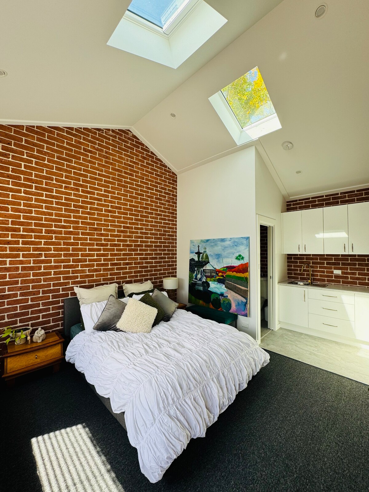 A bright bedroom features a spacious bed adorned with white bedding and decorative pillows. Brick walls add texture, while skylights provide natural light. A colorful artwork and a wooden nightstand are positioned beside the bed, complementing sleek cabinetry in the background.