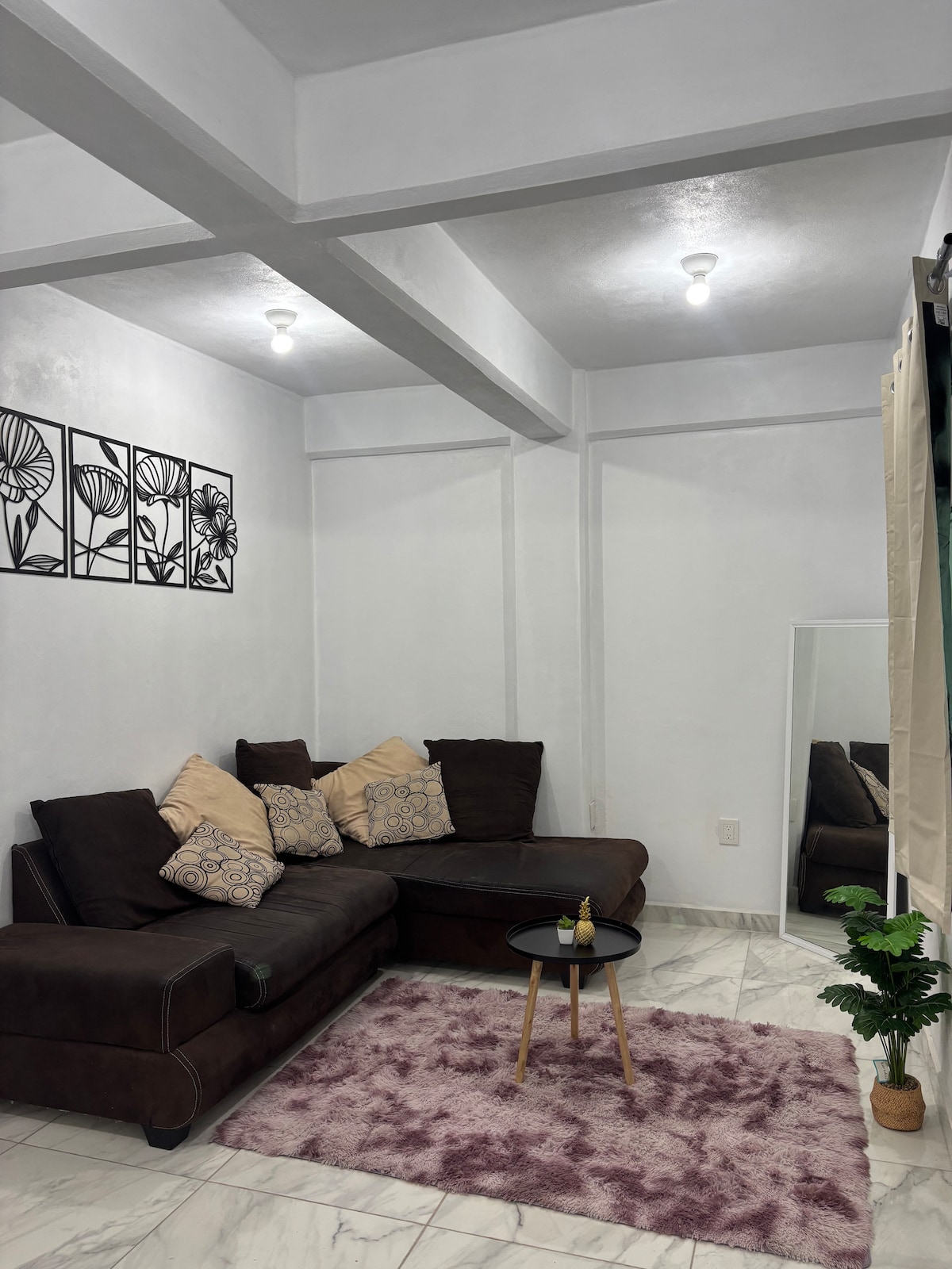 A cozy living area is highlighted by a dark sectional sofa adorned with decorative pillows. A small round table is placed in front of it. A soft rug adds warmth to the space, while a large mirror reflects the bright, neutral-toned walls.