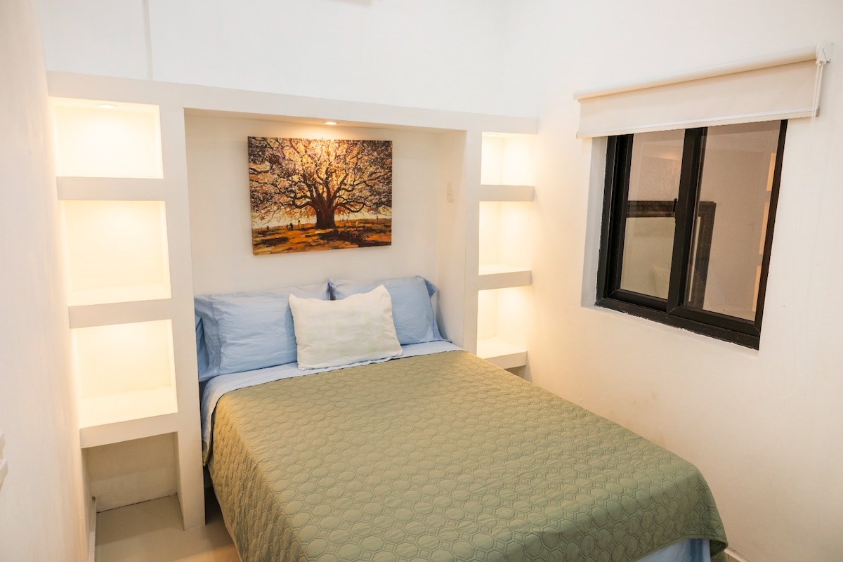 A cozy sleeping area features a spacious bed with a light green coverlet. The walls are painted white, and built-in shelves with soft lighting frame the bed. A painting of a tree adds a serene touch, while a window allows natural light to enter.
