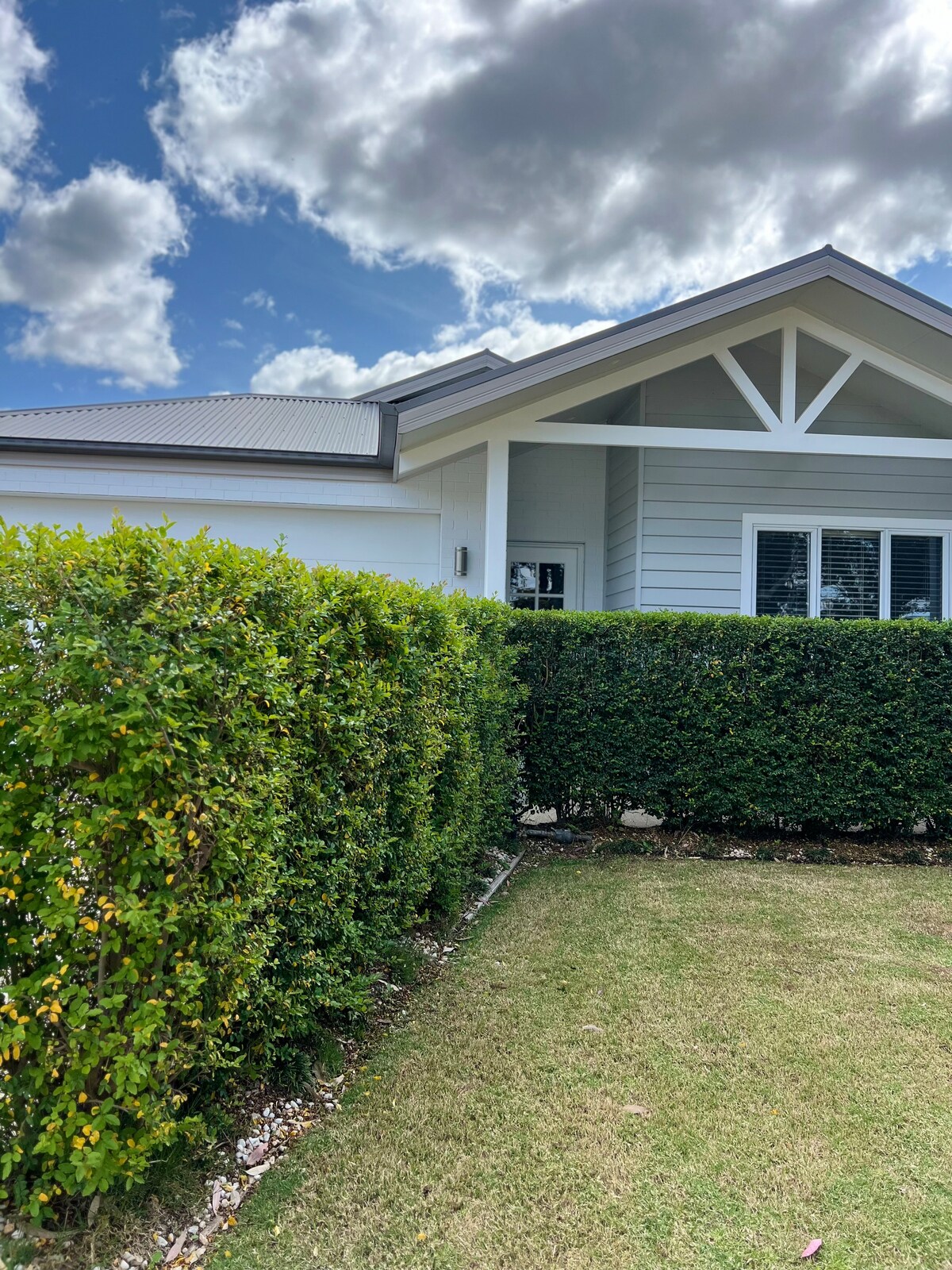 The image presents the exterior of a modern home framed by lush greenery. A well-maintained hedge lines the property, providing privacy. Behind the greenery, architectural details such as a gabled roof and large front windows are visible against a partly cloudy sky.