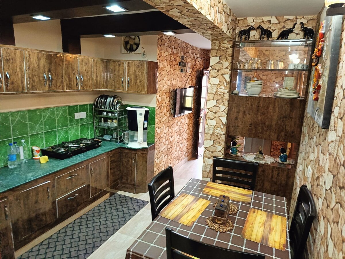 The kitchen area features wooden cabinetry and a green tiled backsplash, with stainless steel cookware displayed neatly. A dining table with dark chairs is positioned nearby. A wall display showcases decorative items, and a sleek kitchen appliance is visible, complementing the warm earthy tones of the decor.