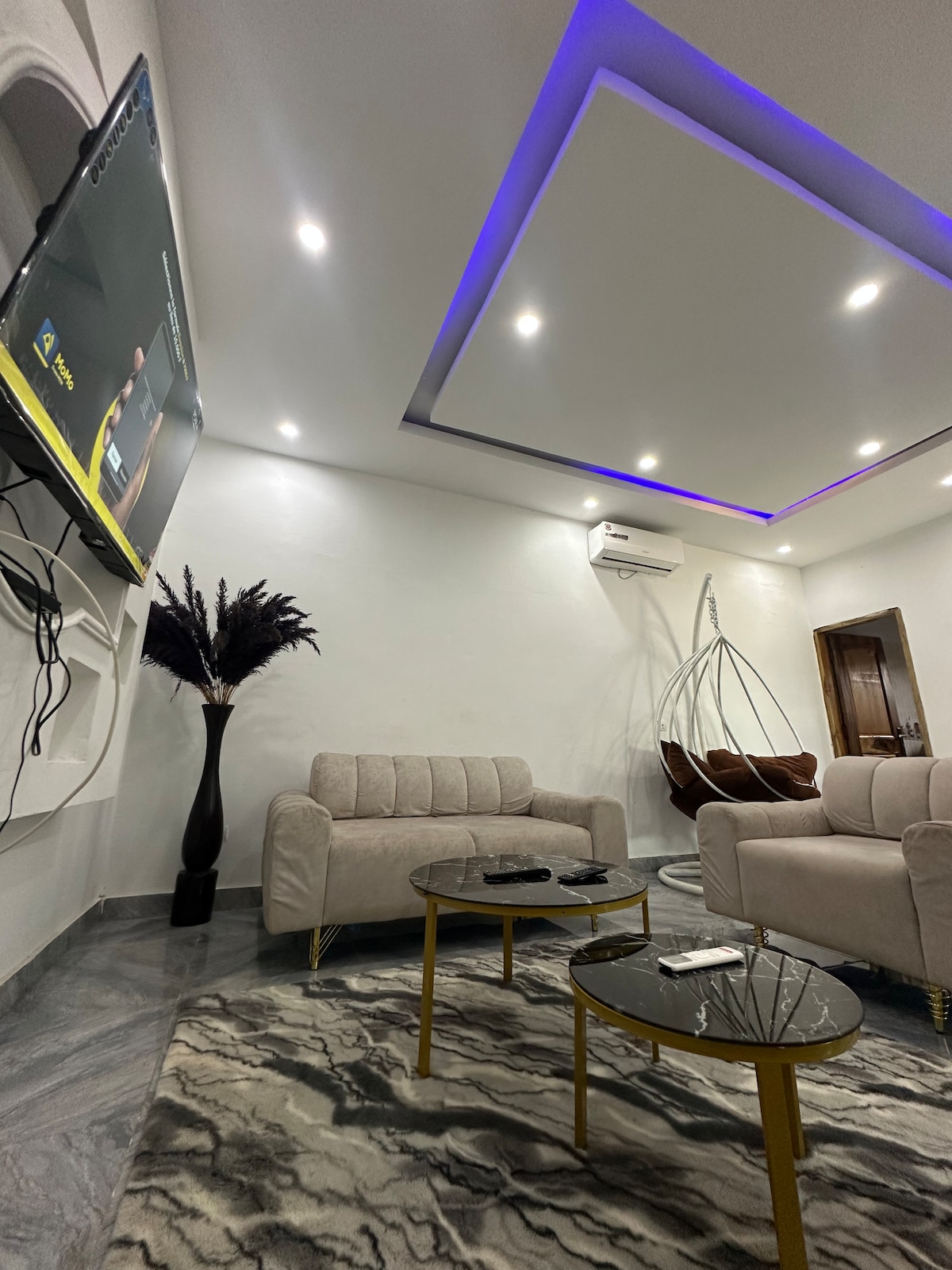 A modern living area is presented with a light-colored sofa and accent chairs. Two glass-top coffee tables rest on a patterned rug. Wall-mounted television and decorative plant elements contribute to the space's ambiance, while recessed lighting highlights the ceiling's design.
