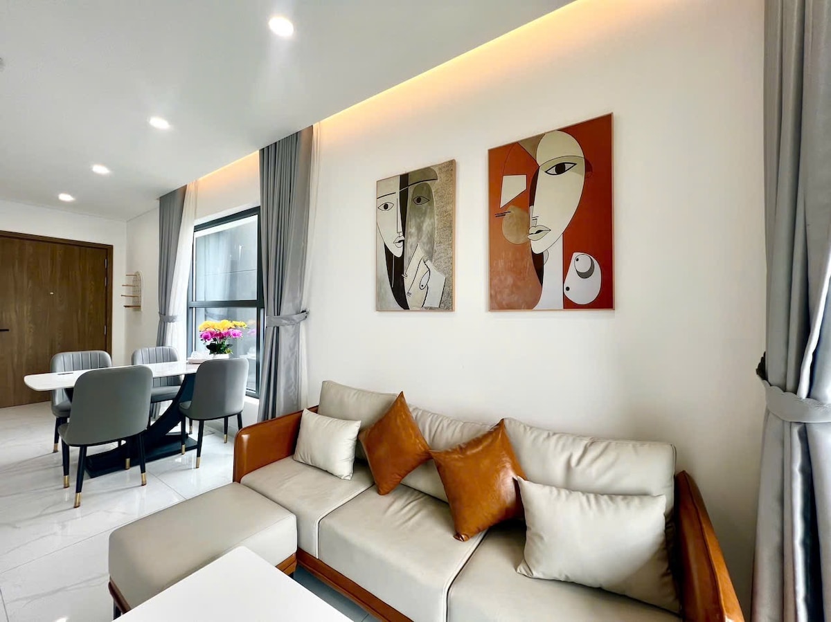 A modern living area features a neutral sofa with contrasting cushions. Abstract wall art adds visual interest, while natural light enters through large windows framed by elegant curtains. A dining table with four chairs is positioned nearby, creating a welcoming space for relaxation and dining.