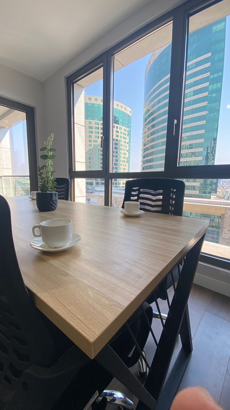 A modern conference room is highlighted by a large wooden table surrounded by black chairs. Two coffee cups are placed on the table, and large windows provide a view of tall, contemporary buildings under clear skies.