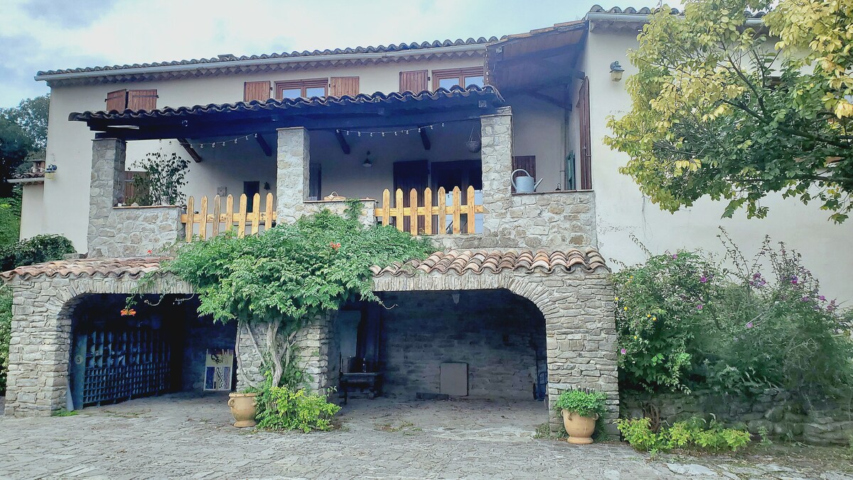 A rustic house is depicted, featuring a stone façade and a charming balcony with wooden railing. Lush greenery surrounds the entrance, complemented by a tiled roof. A spacious courtyard is visible, providing a welcoming area for outdoor relaxation.