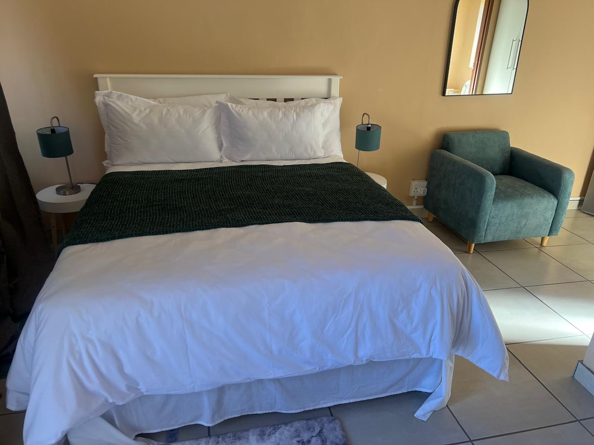 A spacious bed with crisp white linens is positioned against a warm-toned wall. Two bedside lamps emit soft light. A small green armchair complements the room, while a mirror reflects the tranquil ambiance. Tile flooring enhances the serene atmosphere.