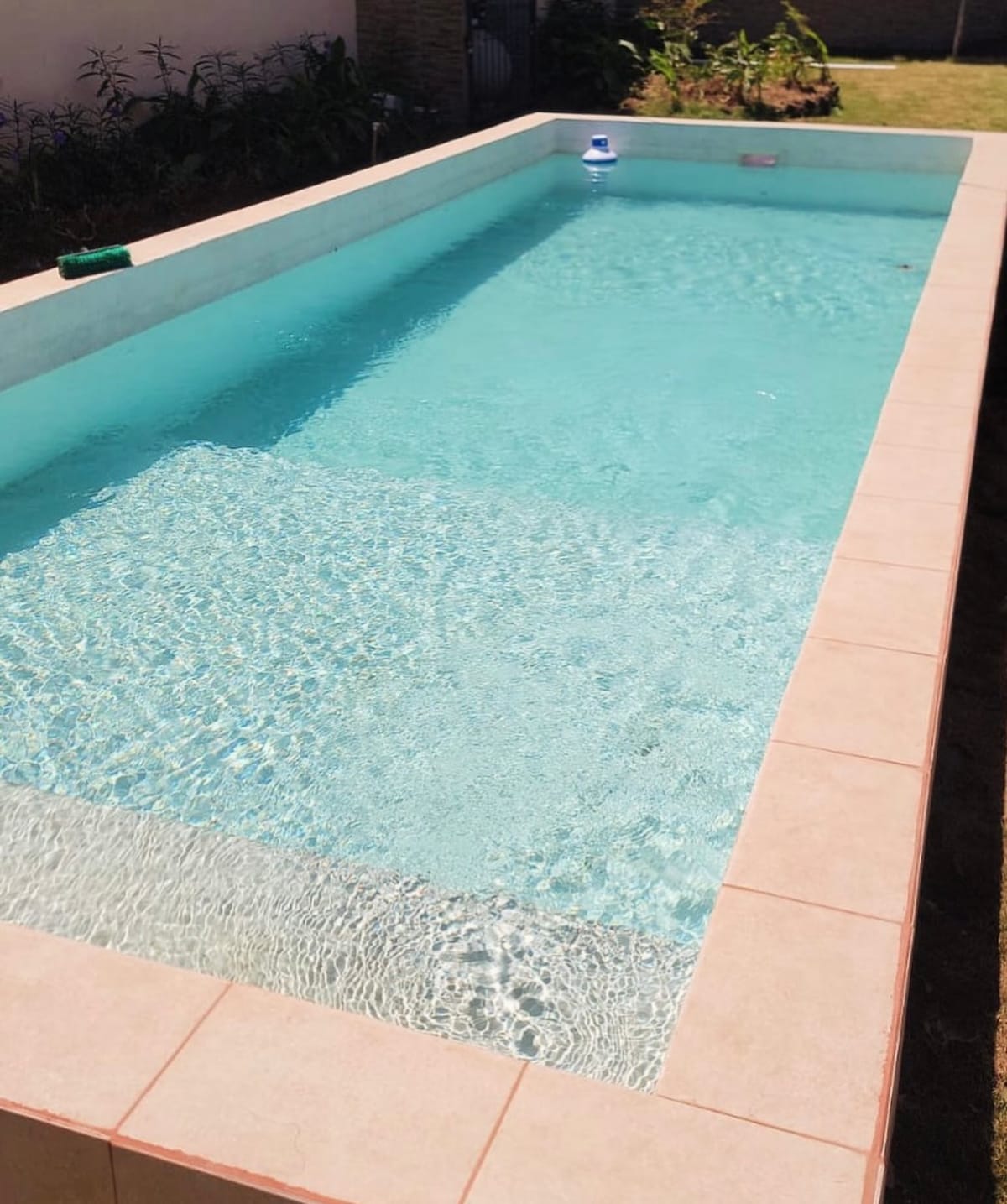 A swimming pool with clear, inviting water is surrounded by light-colored tile. The pool reflects sunlight, creating shimmering patterns on the water's surface. A lawn area is visible alongside the pool, featuring some greenery.