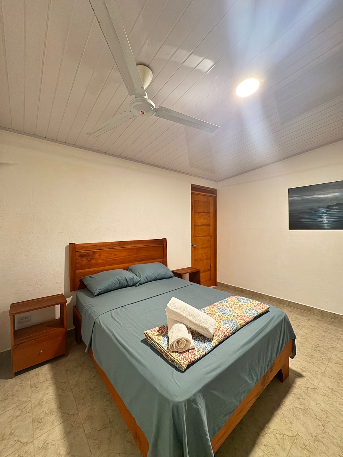 A simple bedroom is featured, furnished with a wooden bed and a matching nightstand. A blanket with a pattern rests atop the bed. A ceiling fan is visible above, while a wood door leads to another space. A serene wall art piece echoes a coastal theme.