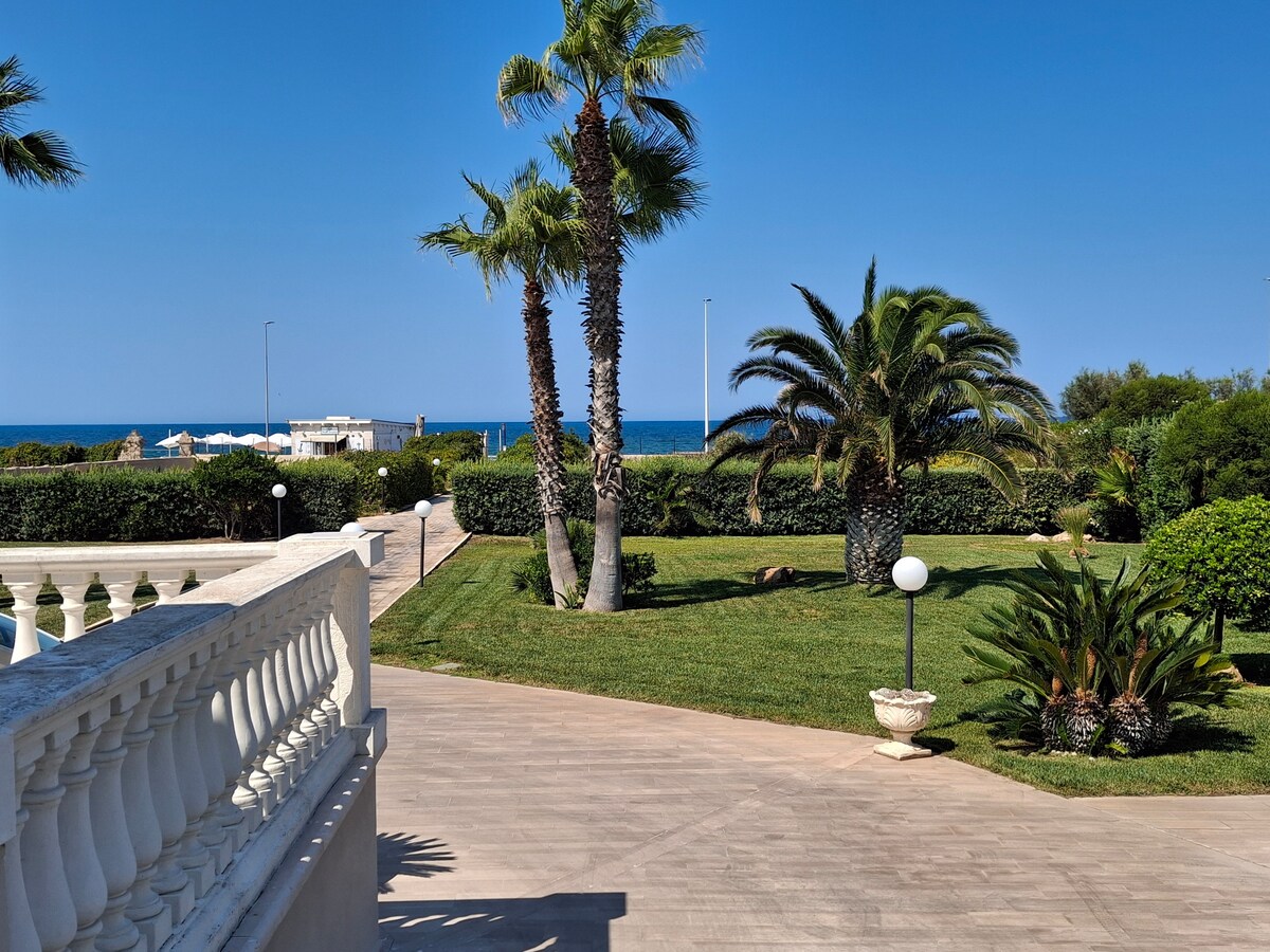 A view from a villa's outdoor steps reveals a well-maintained garden featuring palm trees and lush greenery. The blue Mediterranean sea is visible in the background, contrasting with the bright cloudless sky, creating a serene and inviting environment.