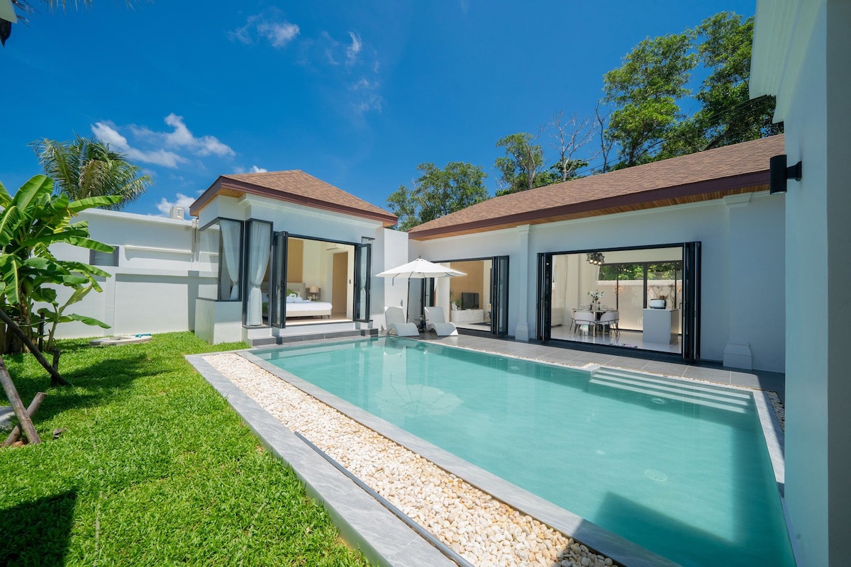 A serene private swimming pool is surrounded by lush green grass, with two comfortable loungers positioned underneath a sun umbrella. The villa's spacious glass doors provide a view into the light-filled living area, revealing an inviting atmosphere perfect for relaxation.