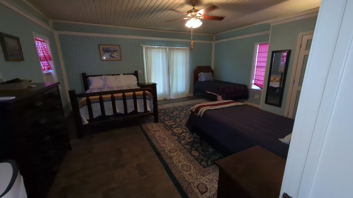 A cozy room features two beds, one with a wooden frame and a spacious mattress. A large window draped in sheer curtains allows natural light to fill the space. Decorative rugs cover the floor, while a mirror and dresser add functional touches.