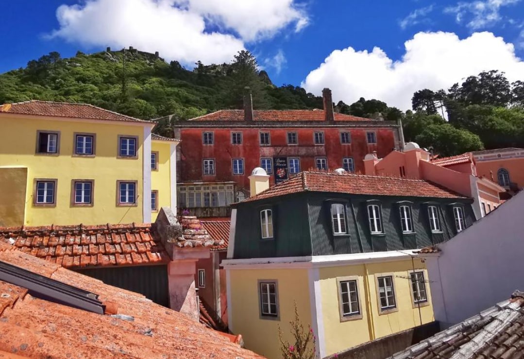 A view of historic buildings with terracotta rooftops showcases a blend of pastel colors against a backdrop of lush greenery and a blue sky. The architectural details of the structures highlight a charming combination of styles, contributing to the scenic landscape of the area.