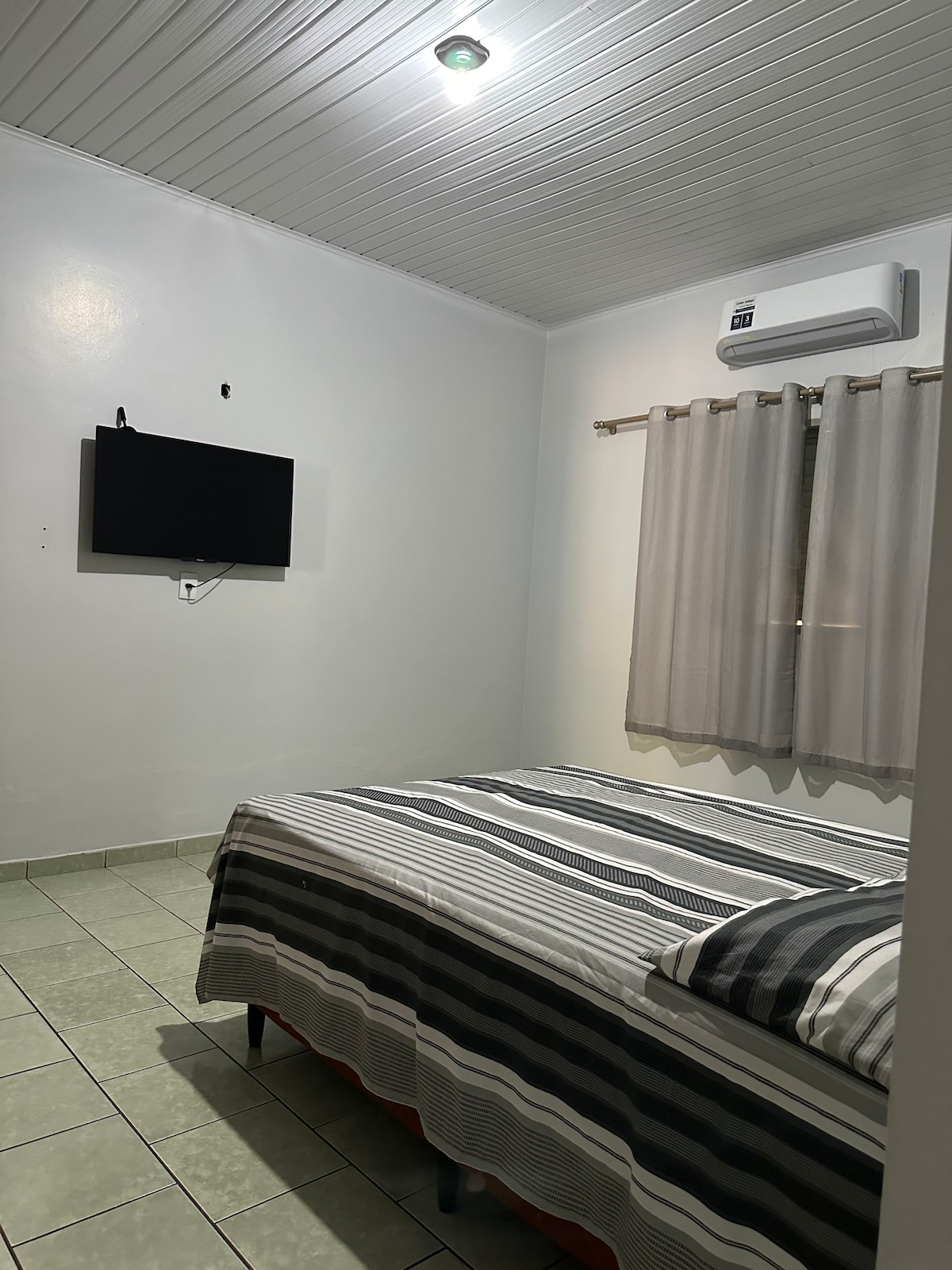 A comfortable bedroom features a spacious bed with a striped blanket. A flat-screen television is mounted on the wall opposite the bed. Soft curtains frame a window, allowing natural light to filter in. A ceiling light provides illumination throughout the room.