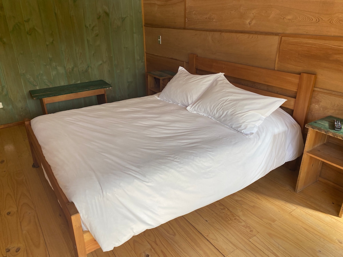 A spacious bed with white linens is positioned against a wooden wall, complemented by natural wood flooring. A small wooden table is located adjacent to the bed, creating a simple and functional sleeping area.