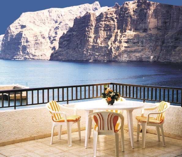 An outdoor terrace features a round table surrounded by four striped chairs. A floral arrangement sits centrally on the table. In the background, the dramatic cliffs of Los Gigantes rise above the ocean, highlighting the stunning coastal landscape.