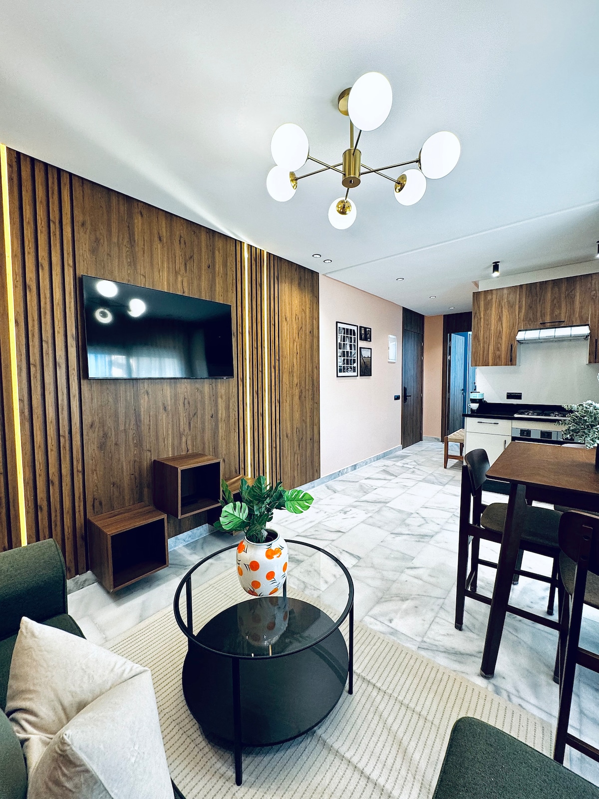 A modern living area is highlighted by wooden paneling and marble flooring. A circular coffee table with a floral centerpiece is positioned centrally, while a mounted television is visible on the wall. The space is illuminated by a stylish light fixture, providing a contemporary touch.