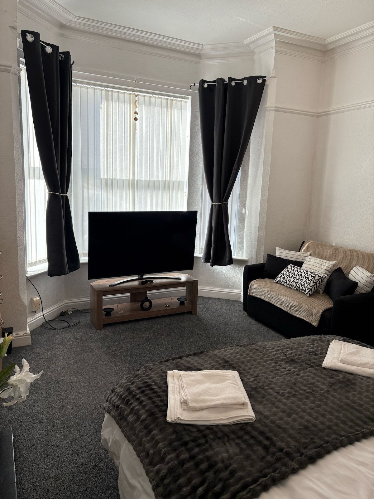A cozy living space features a large television positioned on a wooden stand. Soft furnishings include a black sofa adorned with patterned cushions and a bed with neatly arranged towels. Natural light enters through large windows, complemented by dark curtains.