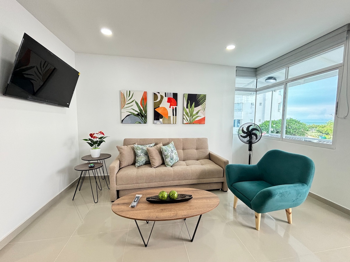 A brightly lit living area features a light brown sofa and a teal accent chair. A round coffee table with a simple design sits in the center, complemented by decorative items. Artwork adorns the wall above the sofa, and large windows allow natural light to fill the space.