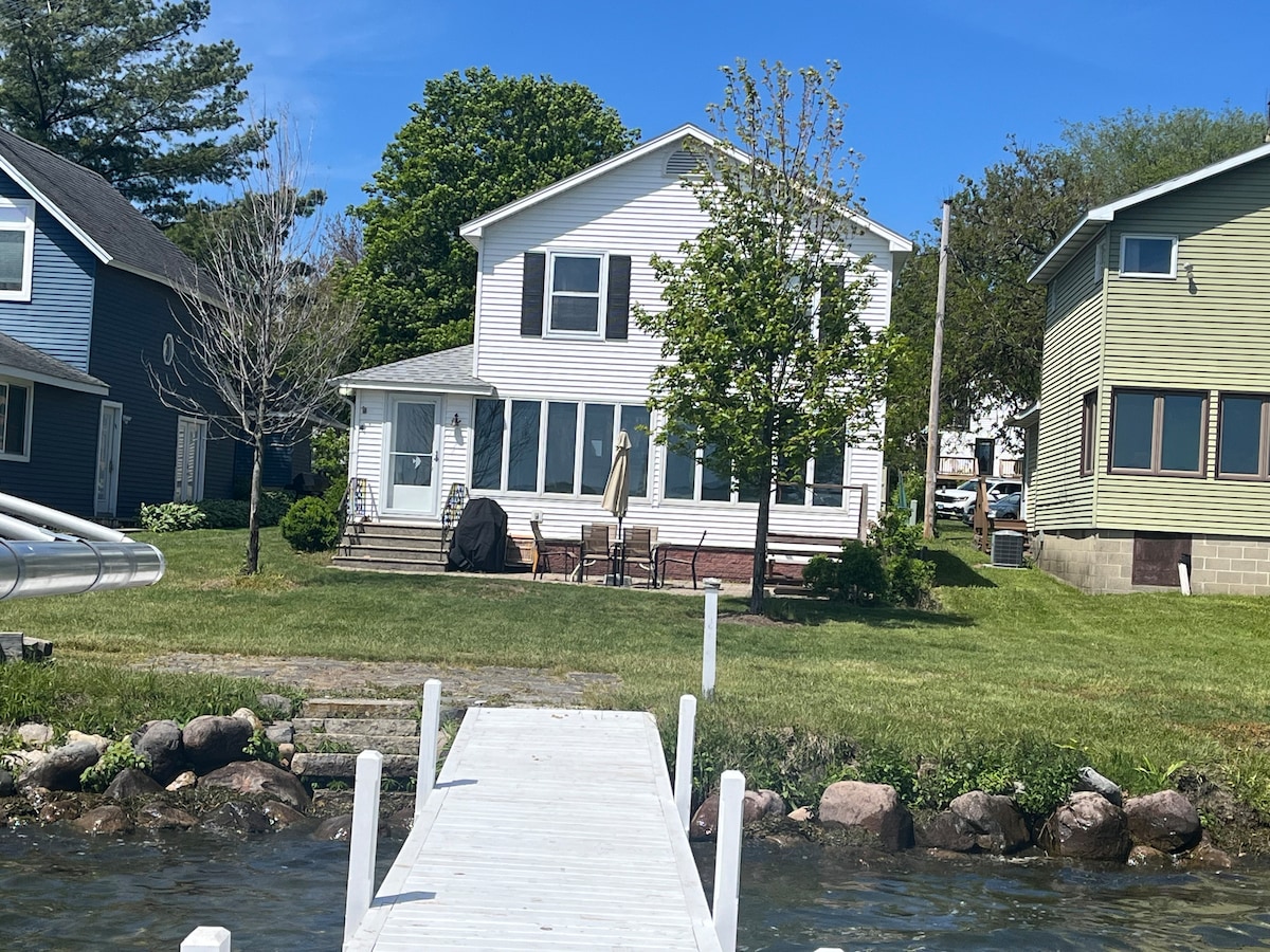 A two-story waterfront home is showcased, with a white exterior and a front porch. The lawn features a centrally-located tree and stone borders, leading to a wooden pier that extends into the water. Nearby homes are visible, enhancing the community feel.
