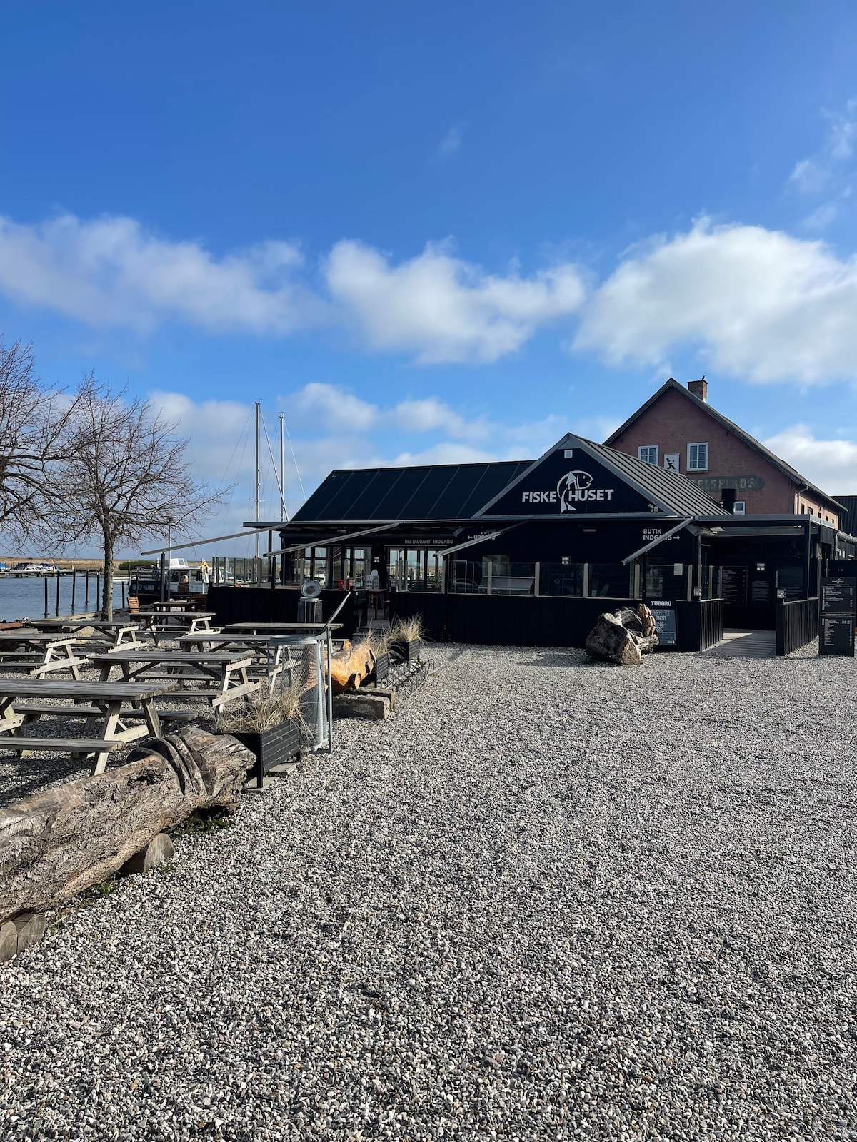 A seafood restaurant is positioned near the water, featuring a black exterior with large windows. Wooden picnic tables are arranged in the gravel area around the building. A few trees are visible in the background, alongside yachts moored in the distance.