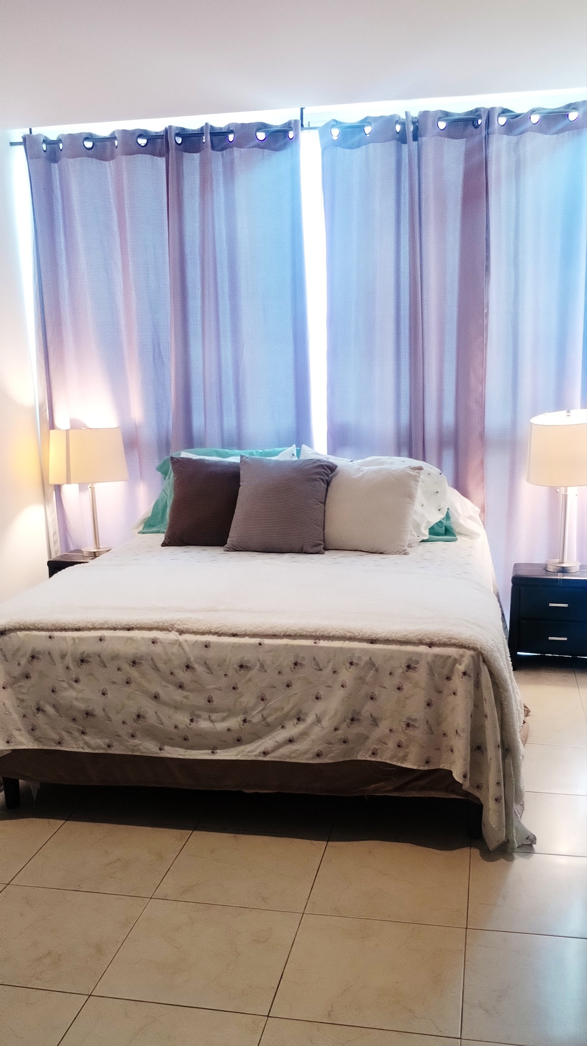 The bedroom features a spacious bed with a light-colored blanket adorned with subtle patterns. Two bedside lamps are positioned on either side, providing additional light. Soft curtains in calming colors frame the window, allowing natural light to gently illuminate the room.