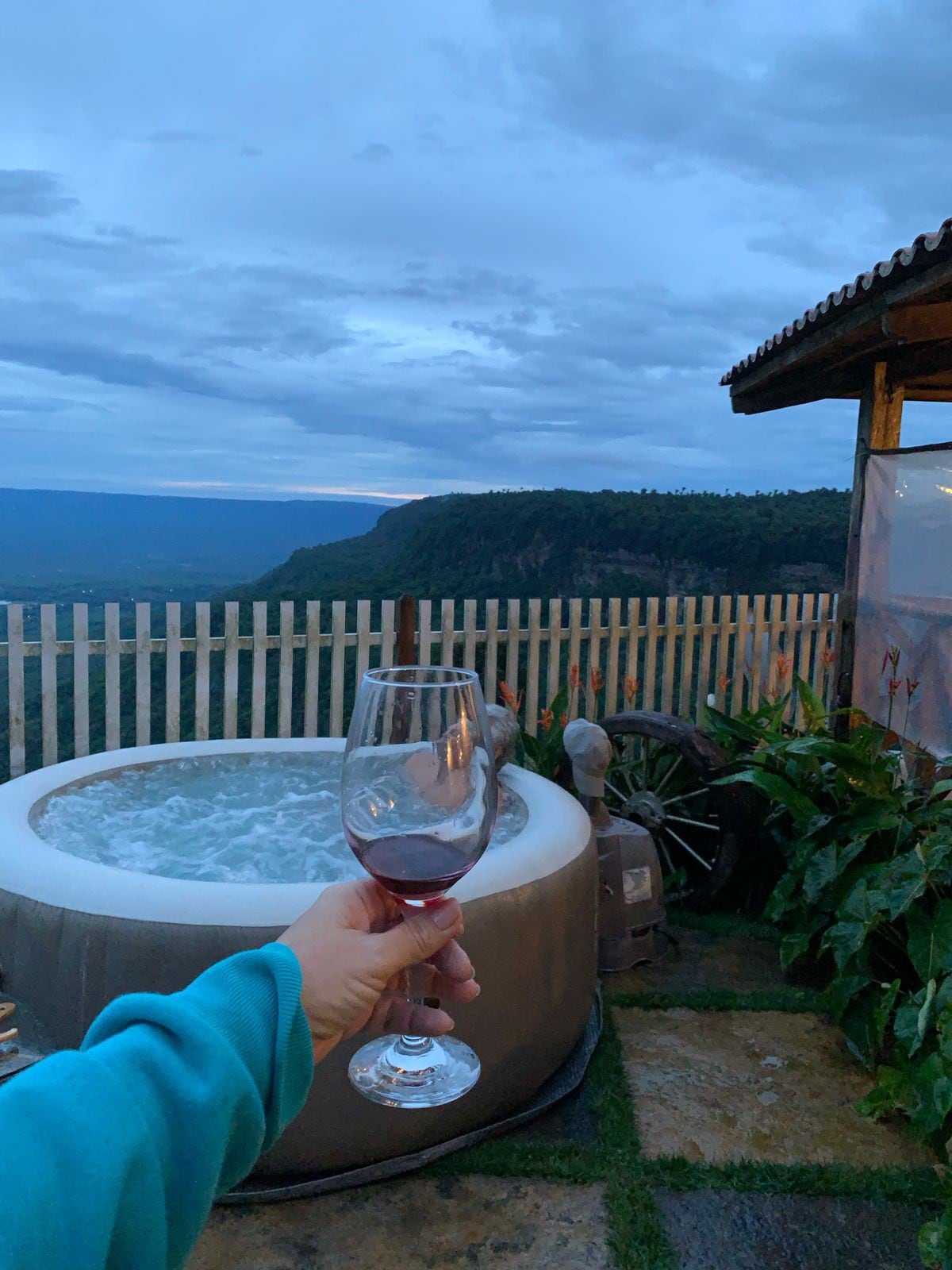 A hot tub is positioned on a patio, surrounded by greenery and a wooden fence. In the foreground, a hand holds a glass of wine. A scenic view of rolling hills and an evening sky is visible in the background.