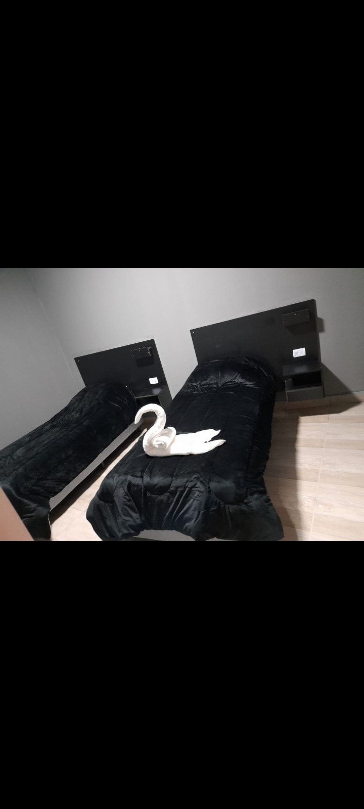 Two twin beds are dressed in black bedding, complemented by dark wooden headboards. A decorative towel swan is positioned on one bed, adding a touch of creativity. The room features gray walls and a light wooden floor, enhancing the overall cozy appearance.