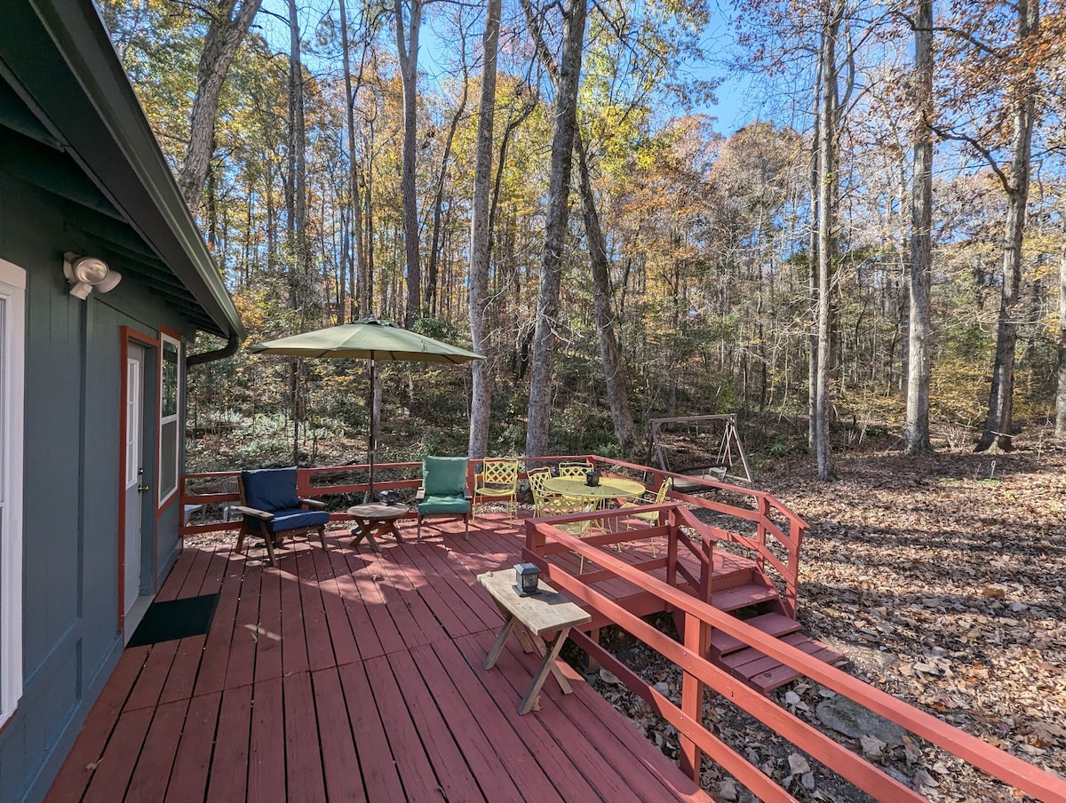 An outdoor deck is surrounded by autumn foliage, featuring outdoor seating with two chairs and a small table. An umbrella provides shade over the seating area. A swing set is visible in the background, set amidst the trees, enhancing the natural surroundings.