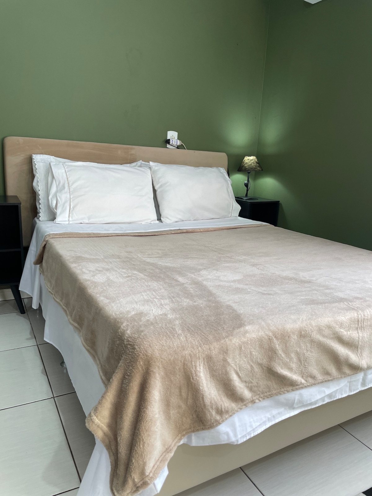 A spacious bed with a soft, beige blanket is positioned against a rich green wall. Two white pillows rest on the headboard. A small lamp on a side table provides gentle lighting, contributing to a relaxed atmosphere.