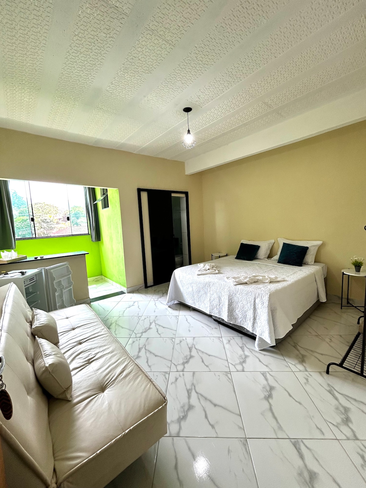 A bright and airy bedroom features a spacious bed dressed in white linens with accent pillows. A cozy leather sofa is positioned nearby, complementing the stylish décor. Natural light fills the room through large windows, showcasing a refreshing view of the outside greenery.