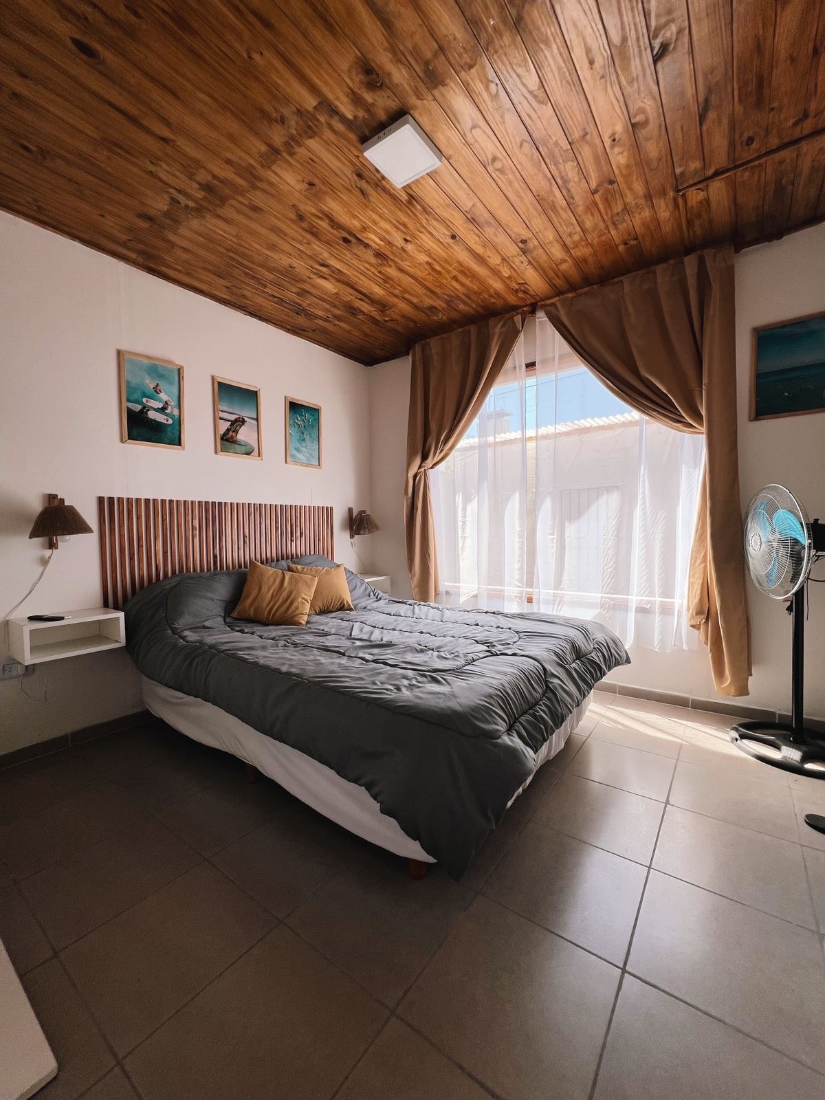A well-lit sleeping area features a spacious bed adorned with a textured duvet and a decorative pillow. Natural light filters through a large window, complemented by soft curtains. Wooden paneling on the ceiling adds warmth, while framed artwork decorates the walls.
