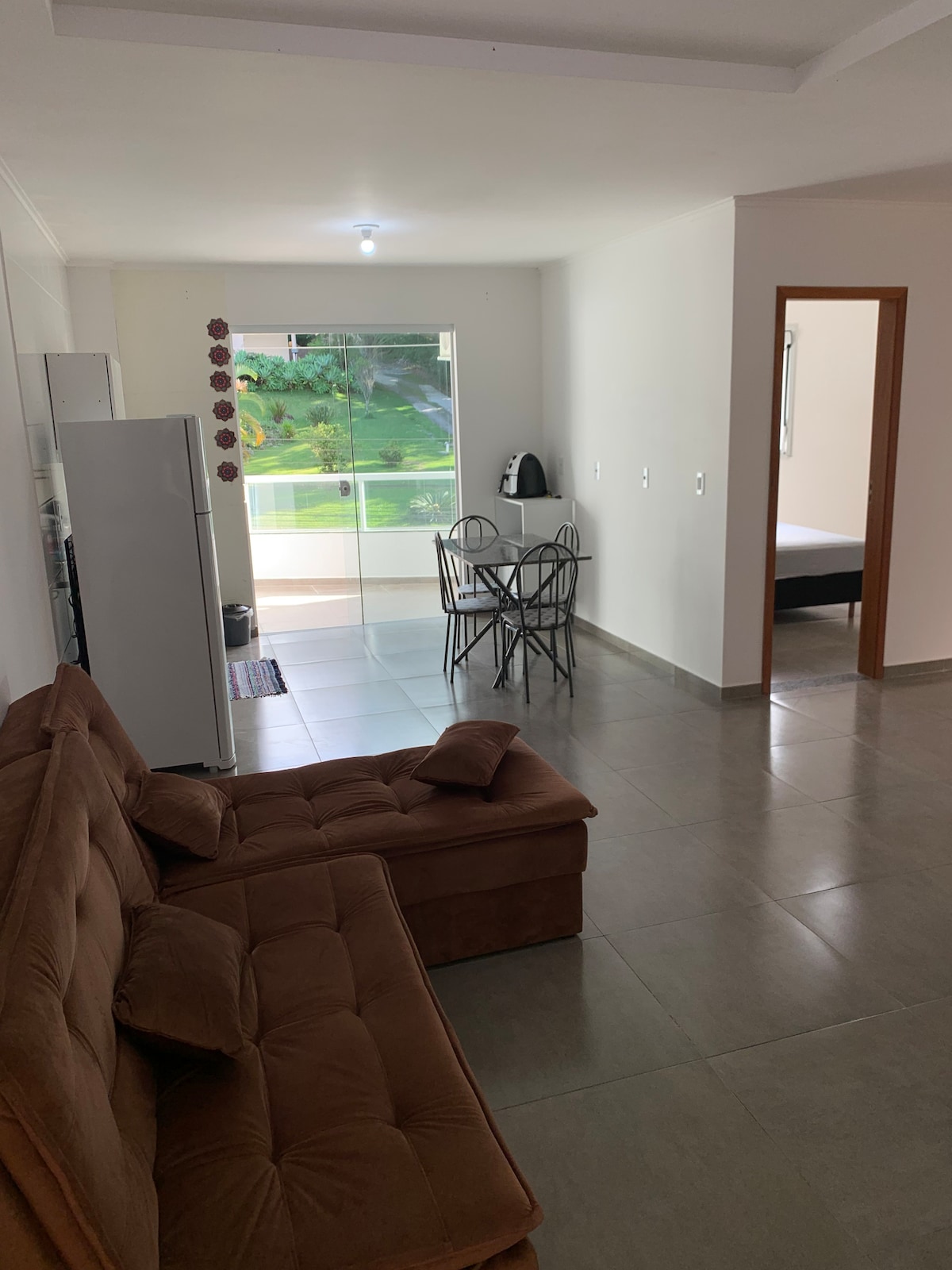 A spacious living area is visible, featuring a comfortable brown sofa and a dining setup with a round table and four chairs. Natural light enters through the large glass doors leading to a balcony, with greenery visible outside. The modern kitchen area includes a refrigerator.