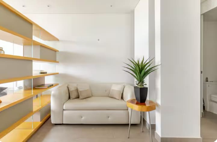 A modern living area features a light-colored sofa with two decorative cushions. A wooden shelf with a warm finish displays various items. A small table with an illuminated plant is positioned beside the sofa, enhancing the overall design of the space.