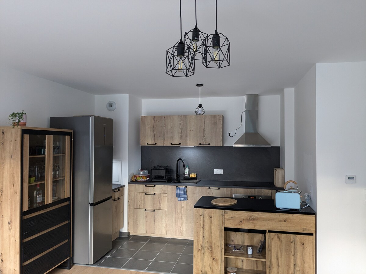 The kitchen is equipped with modern appliances, including a refrigerator, oven, and hood. A wooden cabinetry with a black countertop complements the design. Overhead, three pendant lights provide illumination. The floor is tiled, enhancing the room's functionality and aesthetic appeal.