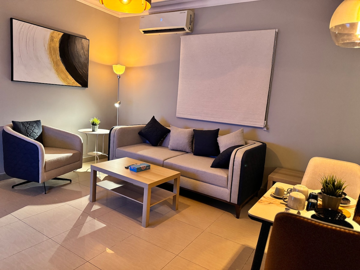 A comfortable living area is showcased with a neutral-toned sofa and two accent chairs. A coffee table is centered, enhancing the space. Soft lighting is provided by a standing lamp and pendant lights, while a large wall art piece adds a modern touch.