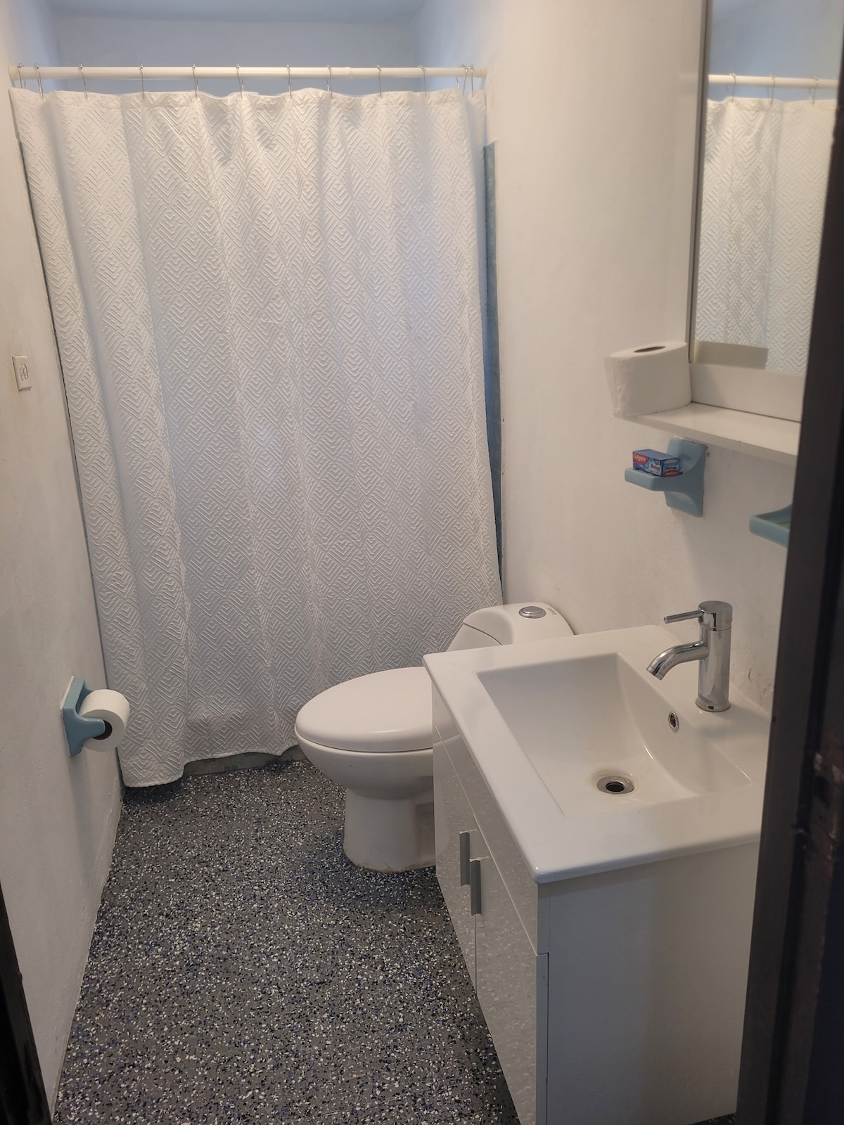 The bathroom features a shower area enclosed by a white curtain, a modern toilet, and a sink with a sleek faucet. The floor is covered with textured gray tiles, and a mirror is mounted above the sink. Essential bathroom supplies are visible for guest convenience.