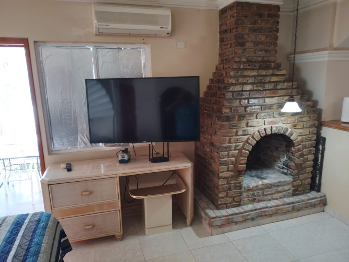 A spacious living area features a large flat-screen television mounted on a cream-colored desk with a matching drawer unit. A traditional brick fireplace stands against the wall, complemented by a modern pendant light hanging above. Natural light enters through a window, creating a bright atmosphere.