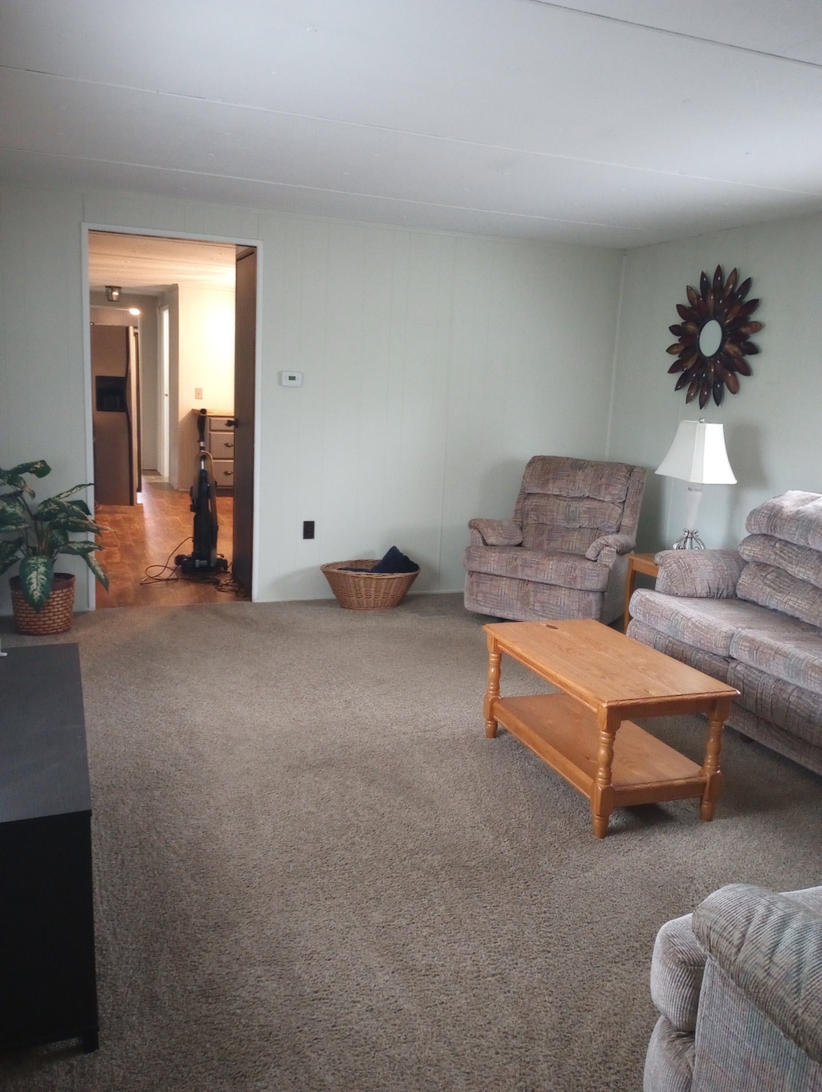 A cozy living area features two comfortable armchairs and a simple coffee table, with a decorative mirror reflecting light on the wall. A small plant brings a touch of greenery, while the space is illuminated by natural light from the adjacent room.